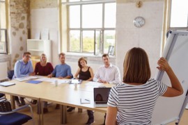 Seminar - Umgang mit Konflikten in der Schule Frau steht neben Flipchart und, Kollegen am Meetingtisch schauen auf Flipchart